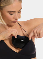 Front view of a model holding Double Sticky Demi Push-Up Pads in black in front of her bra. Demi-shaped silicone bra inserts with smooth outer surface and curved shape visible before placement.