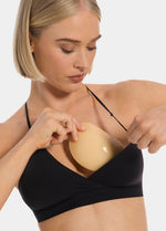 Front view of a model holding Double Sticky Demi Push-Up Pads in latte in front of her bra. Demi-shaped adhesive inserts shown before placement, with smooth outer surface and curved shape visible.